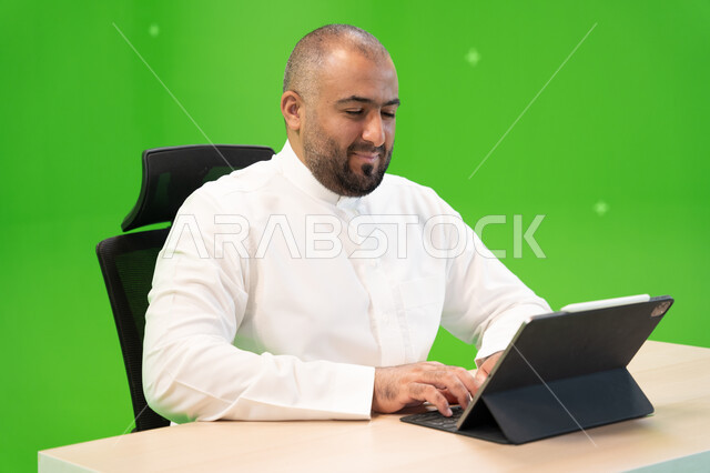 العمل في بيئة مريحة, تحقيق أهداف واستراتيجية الشركة، مفهوم دمج العمل مع التقنية والتكنولوجيا, بورتريه مقرب لرجل عربي سعودي خليجي يرتدي الثوب السعودي التقليدي ينجز مهام العمل باستخدام جهاز التابليت, شركات ووظائف سعودية, خلفية خضراء, كروما.