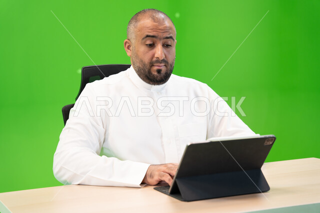 العمل في بيئة مريحة, تحقيق أهداف واستراتيجية الشركة، مفهوم دمج العمل مع التقنية والتكنولوجيا, بورتريه مقرب لرجل عربي سعودي خليجي يرتدي الثوب السعودي التقليدي ينجز مهام العمل باستخدام جهاز التابليت, شركات ووظائف سعودية, خلفية خضراء, كروما.