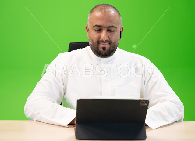 ايماءات تدل على التركيز لتحقيق أهداف الشركة، شركات ووظائف سعودية, إنجاز مهام العمل باستخدام جهاز التابليت, العمل في بيئة مريحة, مفهوم دمج العمل مع التقنية والتكنولوجيا, بورتريه مقرب لرجل خليجي يرتدي الثوب السعودي يضع سماعة البلوتوث، خلفية خضراء, كروما.