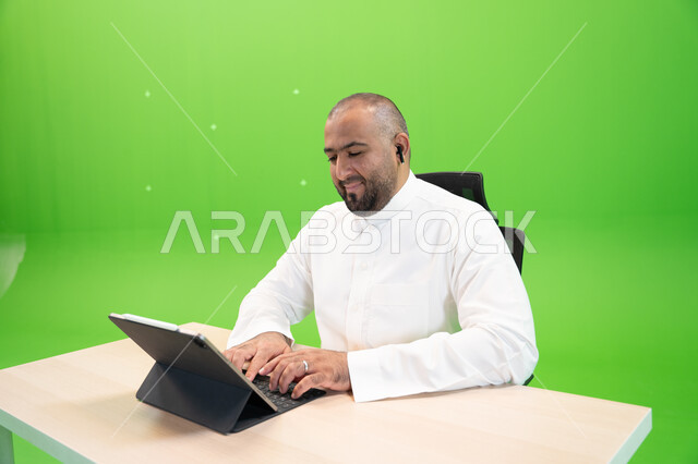 ايماءات تدل على التركيز لتحقيق أهداف الشركة، شركات ووظائف سعودية, إنجاز مهام العمل باستخدام جهاز التابليت, العمل في بيئة مريحة, مفهوم دمج العمل مع التقنية والتكنولوجيا, بورتريه مقرب لرجل خليجي يرتدي الثوب السعودي يضع سماعة البلوتوث، خلفية خضراء, كروما.