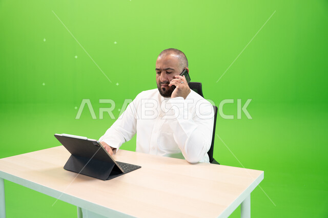  تحقيق استراتيجيات وأهداف العمل، بورتريه مقرب لموظف عربي سعودي خليجي يرتدي الثوب السعودي التقليدي يقوم بإجراء مكالمة هاتفية عبر الهاتف المحمول داخل مكتبه, استخدام جهاز التابليت لإنجاز مهام العمل, إدارة شؤون الأعمال وتنظيمها, خلفية خضراء, كروما.