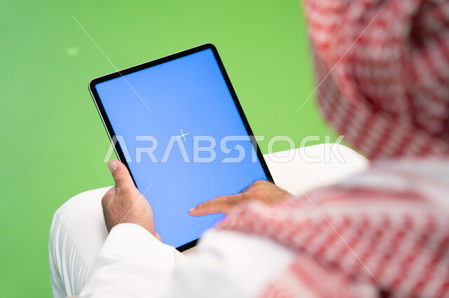 الدردشه والتواصل مع الأقارب والأصدقاء، تصفح مواقع التواصل الاجتماعي عبر الانترنت، بورتريه مقرب من الخلف لرجل عربي خليجي سعودي يرتدي الثوب والشماغ التقليدي يقوم بالنقر على جهاز التابلت بشاشه زرقاء فارغه، التسويق الالكتروني عن بعد، خلفية زرقاء وخضراء، كروما