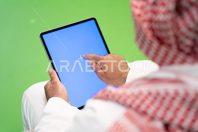 التواصل والدردشه مع الأقارب والأصدقاء، تصفح مواقع التواصل الاجتماعي عبر الانترنت، بورتريه مقرب من الخلف لرجل عربي خليجي سعودي يرتدي الثوب والشماغ التقليدي يقوم بالنقر على جهاز التابلت بشاشه زرقاء فارغه، التسويق الالكتروني عن بعد، خلفية زرقاء وخضراء، كروما