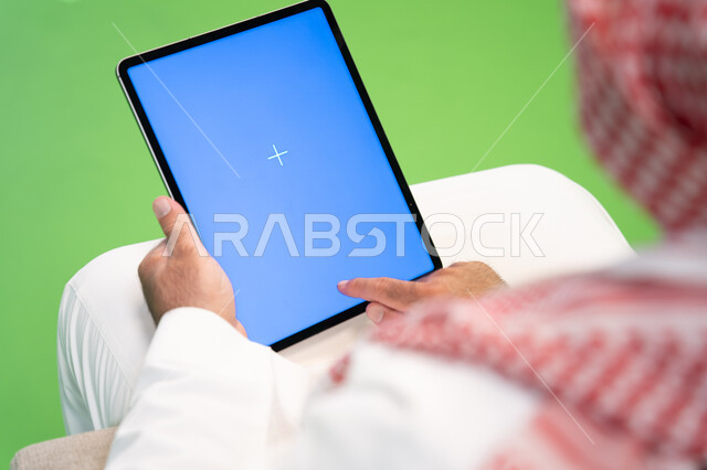 التسويق الالكتروني عن بعد، تصفح مواقع التواصل الاجتماعي عبر الانترنت، بورتريه مقرب من الخلف لرجل عربي خليجي سعودي يرتدي الثوب والشماغ التقليدي يقوم بالنقر على جهاز التابلت بشاشه زرقاء فارغه، التواصل والدردشه مع الأقارب والأصدقاء، خلفية زرقاء وخضراء، كروما
