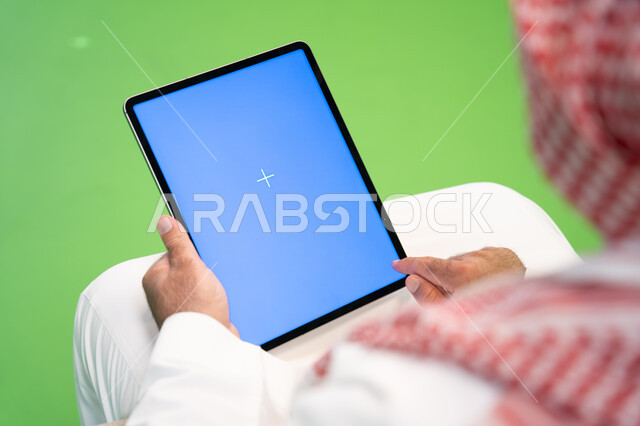Browsing social networking sites online, a close-up portrait from the back of a Saudi Gulf Arab man wearing a traditional thobe and shemagh, tapping on a tablet with a blank blue screen, communicating and chatting with relatives and friends, remote electr