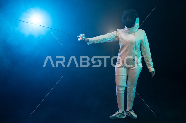 التدرب على الحركات بسيف المبارزة، فنون القتال الفردية، ممارسة رياضة مبارزة السلاح النسائية، لاعبة مبارزة عربية خليجية سعودية ترتدي زي المسايفة وتحمل بيدها سيف، رياضات المسايفة الأولومبية، صورة الجسم بالطول الكامل، خلفية سوداء مع انارة زرقاء