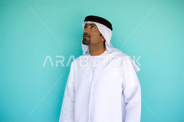 النظر الى شيء ما في الأعلى, الاهتمام بالمظهر الخارجي, الوقوف باستقامة وثقة, بورتريه  لرجل عربي خليجي امارتي يرتدي الكندورة والغترة الاماراتية , خلفية زرقاء