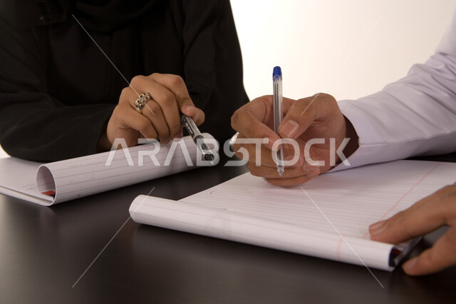 إجراء المناقشات وتنظيم الأعمال, الشراكة والتعاون في انجاز العمل, إعداد الخطط الاستراتيجية, صورة مقربة ليد رجل عربي خليجي اماراتي يرتدي الكندورة الاماراتية و يد رجل بالبدلة الرسمية في غرفة الإجتماعات يقومان بتدوين الملاحظات والأفكار علبى دفتر الأعمال