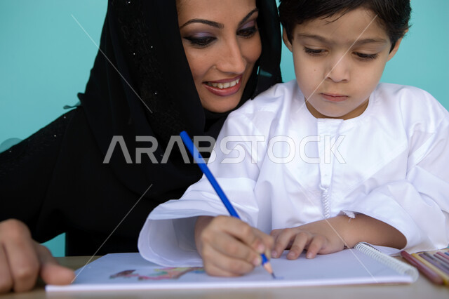 أم تساعد ابنها على الرسم بايماءات وجه تدل على السعادة والفخر، رعاية وتنمية مهارات الأطفال، بورتريه مقرب لطفل عربي خليجي إماراتي يرتدي الزي الإماراتي يقوم بالرسم والتلوين رفقة والدته، التعلم والابداع وتنمية المهارات، خلفية زرقاء
