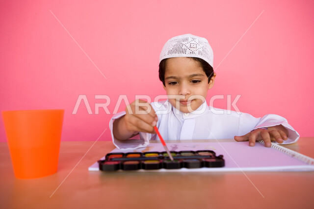 مفهوم الاهتمام بالهوايات, إيماءات تدل على التركيز والاستمتاع بالرسم و التلوين، تنمية المهارات الفنية في الروضات الإماراتية، بورتريه لطفل عربي خليجي إماراتي يرتدي الزي الإماراتي التقليدي يقوم بالرسم والتلوين بالألوان المائية, خلفية وردية