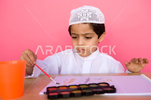 إيماءات تدل على التركيز والاستمتاع بالرسم و التلوين، تنمية المهارات الفنية في الروضات الإماراتية، بورتريه لطفل عربي خليجي إماراتي يرتدي الزي الإماراتي التقليدي يقوم بالرسم والتلوين بالألوان المائية, مفهوم الاهتمام بالهوايات, خلفية وردية