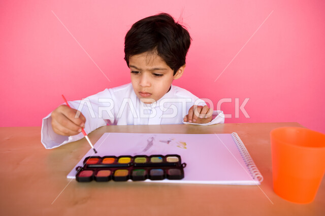 إيماءات تدل على التركيز والاستمتاع بالرسم و التلوين، تنمية المهارات الفنية في الروضات الإماراتية، بورتريه لطفل عربي خليجي إماراتي يرتدي الزي الإماراتي التقليدي يقوم بالرسم والتلوين بالألوان المائية, مفهوم الاهتمام بالهوايات, خلفية وردية