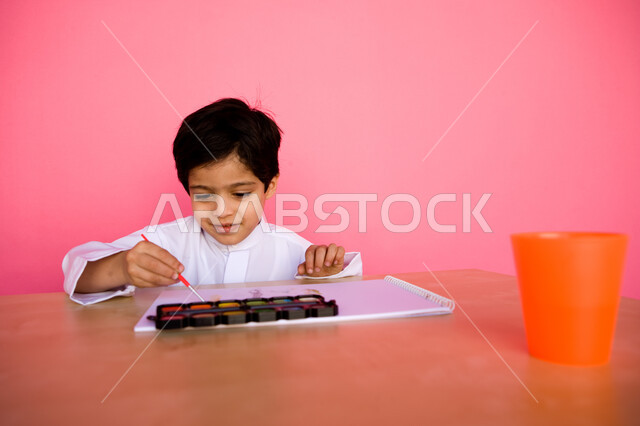 إيماءات تدل على التركيز والاستمتاع بالرسم و التلوين، تنمية المهارات الفنية في الروضات الإماراتية، بورتريه لطفل عربي خليجي إماراتي يرتدي الزي الإماراتي التقليدي يقوم بالرسم والتلوين بالألوان المائية, مفهوم الاهتمام بالهوايات, خلفية وردية