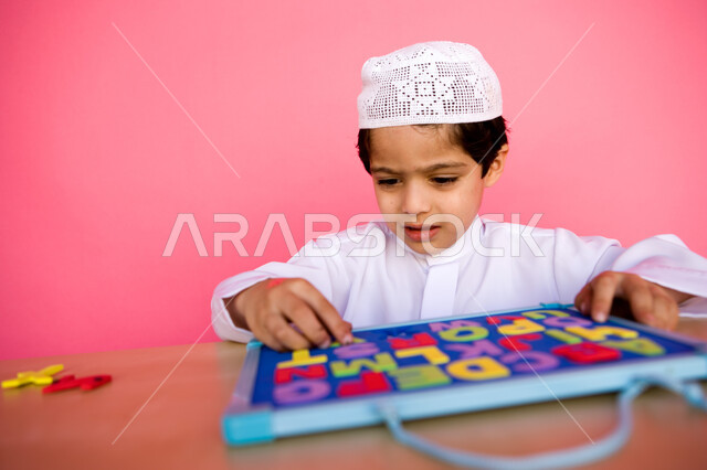 مهارة التعلم عن طريق الألعاب التعليمية، استخدام أشكال الحروف الانكليزية والتعرف عليها، بورتريه مقرب لطفل عربي خليجي إماراتي يرتدي الزي الاماراتي ويلعب بلعبة لغز الأبجدية , الاستمتاع بالنشاطات الترفيهية المفيدة, خلفية ملونة