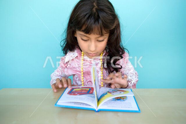 تنمية مهارات القراءة للأطفال, إيماءات الانتباه والتركيز بالمطالعة, بورتريه مقرب لفتاة عربية خليجية إماراتية تقوم بالقراءة بتمعن , مطالعة القصص التصويرية المسلية, تمضية أوقات الفراغ بالتعلم والتثقف الذاتي, الاهتمام بتصفح الكتب المفيدة, خلفية زرقاء