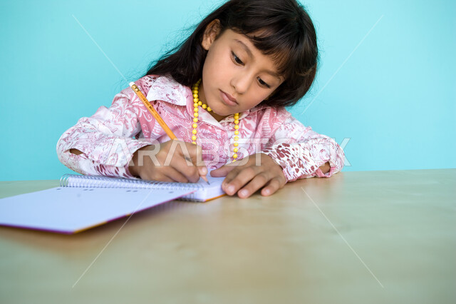 Integration and focus on taking notes and daily assignments, performing school tasks at home, close-up portrait of an Arab Gulf Emirati girl writing on a notebook, developing writing skills since childhood, blue background