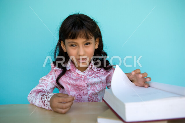 الاهتمام بتصفح الكتب المفيدة, شغف التعرف على الكتب المحتلفة, بورتريه مقرب لفتاة عربية خليجية إماراتية تقوم بتقليب صفحات الكتاب وتنظر الى الكاميرا , حب المطالعة و الروايات, تمضية وقت الفراغ بالتعلم والتثقف الذاتي, تنمية مهارات القراءة للأطفال, خلفية زرقاء