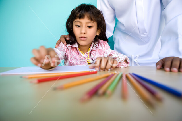 رعاية المواهب والاهتمام بها , دور الأهل في تطوير وتنمية المهارات, إيماءات السعادة والاستمتاع بمشاركة الأنشطة الفنية مع الأب، شغف وحب الرسم، بورتريه مقرب لفتاة صغيرة عربية خليجية إماراتية تقوم بالرسم والتلوين بالأقلام الخشبية رفقة والدها، خلفية زرقاء