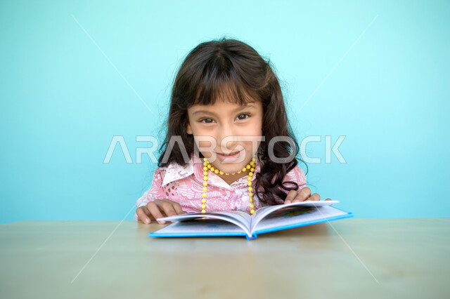 تنمية مهارات القراءة للأطفال, النظر الى الكاميرا, بورتريه مقرب لفتاة عربية خليجية إماراتية تقوم بالقراءة بتمعن , مطالعة القصص التصويرية المسلية, تمضية أوقات الفراغ بالتعلم والتثقف الذاتي, الاهتمام بتصفح الكتب المفيدة, خلفية زرقاء