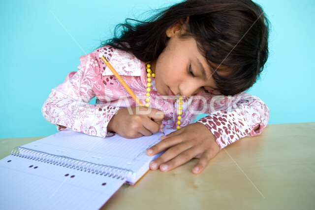 الاندماج والتركيز على تدوين الملاحظات والواجبات اليومية, أداء الوظائف المدرسية في المنزل, بورتريه مقرب لفتاة عربية خليجية إماراتية تقوم بالكتابة على دفتر الملاحظات, تطوير مهارات الكتابة منذ الصغر, خلفية زرقاء