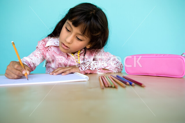 أدوات وقرطاسية التلوين، تطوير وتنمية الهوايات, إيماءات الاندماج والتركيز في الأنشطة الفنية، بورتريه مقرب لفتاة صغيرة عربية خليجية إماراتية تقوم بالرسم والتلوين بالأقلام الخشبية، رعاية المواهب والاهتمام بها في الإمارات العربية المتحدة, خلفية زرقاء