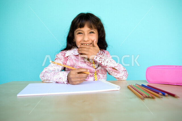  أدوات وقرطاسية التلوين، تطوير وتنمية الهوايات, إيماءات الاستمتاع بالأنشطة الفنية، بورتريه مقرب لفتاة صغيرة عربية خليجية إماراتية تقوم بالابتسام و الضحك وترسم بقلم الرصاص، رعاية المواهب والاهتمام بها في الإمارات العربية المتحدة, خلفية زرقاء