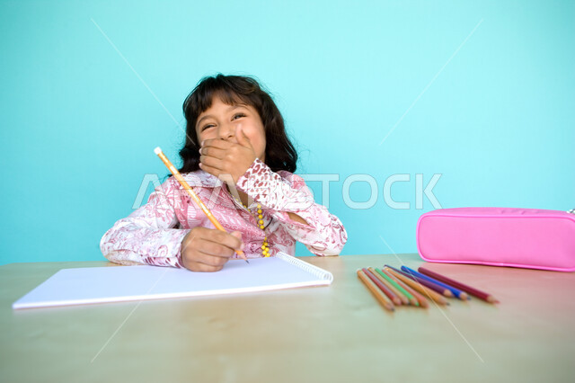  أدوات وقرطاسية التلوين، تطوير وتنمية الهوايات, إيماءات الاستمتاع بالأنشطة الفنية، بورتريه مقرب لفتاة صغيرة عربية خليجية إماراتية تقوم بالابتسام و الضحك وترسم بقلم الرصاص، رعاية المواهب والاهتمام بها في الإمارات العربية المتحدة, خلفية زرقاء