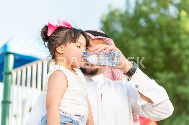 فتاه جميلة مصابة بملازمة داون تقضي وقتها في نزهه مع والدها بالمنتزه ، اب سعودي خليجي يساعد ابنته على شرب الماء ، أصحاب الهمم ، ذوي الاحتياجات الخاصة