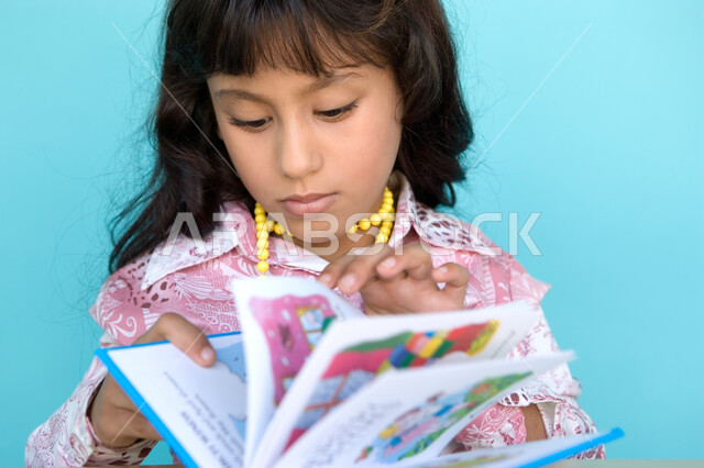 تمضية أوقات الفراغ بالتعلم والتثقف الذاتي, إيماءات الانتباه والتركيز بالمطالعة, بورتريه مقرب لفتاة عربية خليجية إماراتية تقوم بالقراءة بتمعن , مطالعة القصص التصويرية المسلية, تنمية مهارات القراءة للأطفال, الاهتمام بتصفح الكتب المفيدة, خلفية زرقاء