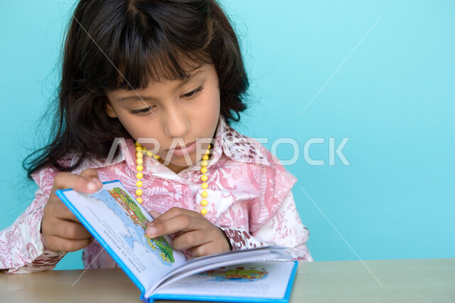 مطالعة القصص التصويرية المسلية, إيماءات الانتباه والتركيز بالمطالعة, بورتريه مقرب لفتاة عربية خليجية إماراتية تقوم بالقراءة بتمعن ,  تمضية أوقات الفراغ بالتعلم والتثقف الذاتي, تنمية مهارات القراءة للأطفال, الاهتمام بتصفح الكتب المفيدة, خلفية زرقاء