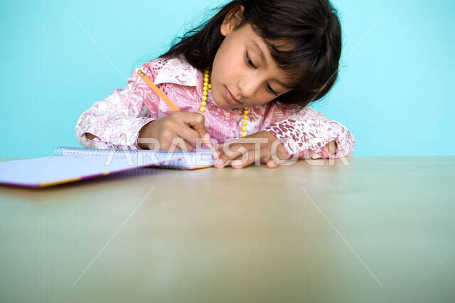 Integration and focus on taking notes and daily assignments, performing school tasks at home, close-up portrait of an Arab Gulf Emirati girl writing on a notebook, developing writing skills since childhood, blue background