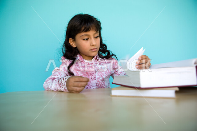 شغف التعرف على الكتب المحتلفة, بورتريه مقرب لفتاة عربية خليجية إماراتية تقوم بتقليب صفحات الكتاب , حب المطالعة و الروايات, تمضية أوقات الفراغ بالتعلم والتثقف الذاتي, تنمية مهارات القراءة للأطفال, الاهتمام بتصفح الكتب المفيدة, خلفية زرقاء