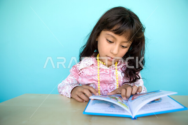 Gestures of attention and focus while reading, a close-up portrait of an Arab Gulf Emirati girl reading carefully, reading entertaining pictorial stories, spending free time with learning and self-education, developing children’s reading skills, interest 