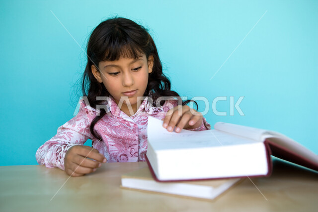 شغف التعرف على الكتب المحتلفة, بورتريه مقرب لفتاة عربية خليجية إماراتية تقوم بتقليب صفحات الكتاب , حب المطالعة و الروايات, تمضية أوقات الفراغ بالتعلم والتثقف الذاتي, تنمية مهارات القراءة للأطفال, الاهتمام بتصفح الكتب المفيدة, خلفية زرقاء