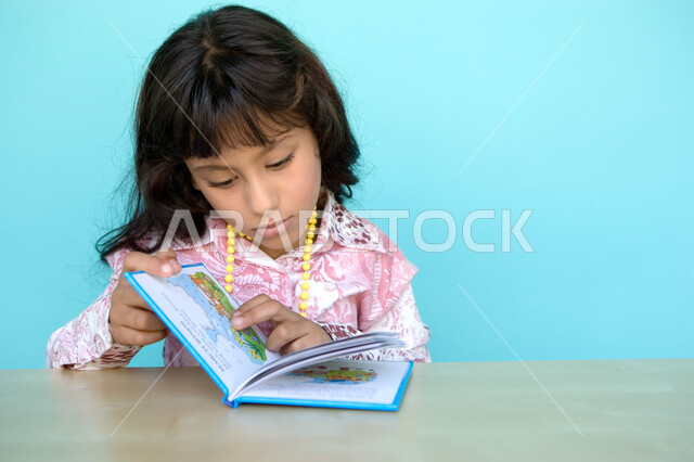 Gestures of attention and focus while reading, a close-up portrait of an Arab Gulf Emirati girl reading carefully, reading entertaining pictorial stories, spending free time with learning and self-education, developing children’s reading skills, interest 