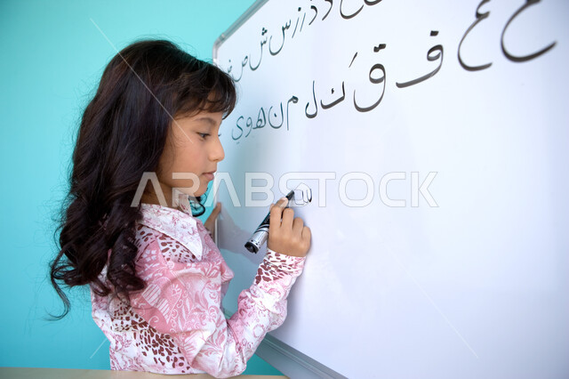 تعليم التلاميذ تكوين الكلمات باللغة العربية، تنمية القدرات اللغوية والفكرية للطلاب, بورتريه مقرب من الجانب لفتاة عربية خليجية إماراتية تحمل بيدها قلم اللوح و تقوم بكتابة الكلمات على السبورة, متعة حصة اللغة العربية, خلفية زرقاء