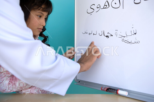 جودة التعليم في الإمارات، تنمية القدرات اللغوية والفكرية للطلاب, تعليم التلاميذ تكوين الكلمات باللغة العربية، بورتريه مقرب من الجانب لأستاذ عربي خليجي إماراتي يقوم بتعليم طالبة تشكيل الجمل في حصة اللغة العربية على السبورة, خلفية زرقاء