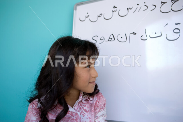 جودة التعليم في الإمارات، تنمية القدرات اللغوية والفكرية للطلاب, تعليم التلاميذ تكوين الكلمات باللغة العربية، بورتريه مقرب من الجانب لفتاة عربية خليجية إماراتية تقف أمام اللوح وتنظر الى الحروف العربية, متعة حصة اللغة العربية, خلفية زرقاء