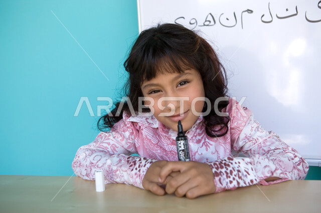 الاستمتاع بحصة اللغة العربية, تنمية القدرات اللغوية والفكرية للطلاب, تعليم التلاميذ تكوين الكلمات باللغة العربية، بورتريه مقرب لفتاة عربية خليجية إماراتية مبتسمة تنظر الى الكاميرا و تحمل بيدها قلم اللوح, خلفية زرقاء