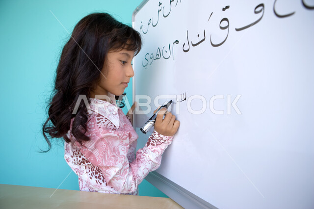 تعليم التلاميذ تكوين الكلمات باللغة العربية، تنمية القدرات اللغوية والفكرية للطلاب, بورتريه مقرب من الجانب لفتاة عربية خليجية إماراتية تحمل بيدها قلم اللوح و تقوم بكتابة الكلمات على السبورة, متعة حصة اللغة العربية, خلفية زرقاء
