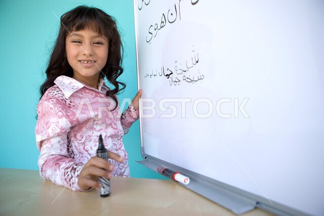 الوقوف أمام السبورة, متعة حصة اللغة العربية, تنمية القدرات اللغوية والفكرية للطلاب, تعليم التلاميذ تكوين الكلمات باللغة العربية، بورتريه مقرب من الجانب لفتاة عربية خليجية إماراتية مبتسمة تنظر الى الكاميرا  و تحمل بيدها قلم اللوح , خلفية زرقاء