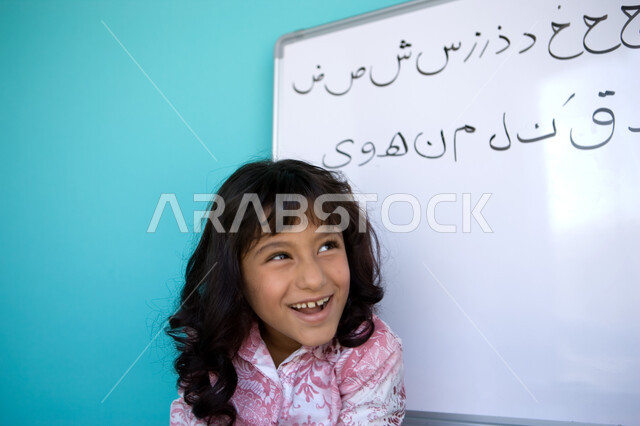 Developing students’ linguistic skills, teaching students to form words in Arabic, a close-up portrait of an Arab Gulf Emirati girl laughing and smiling and standing in front of the blackboard, fun Arabic language class, blue background