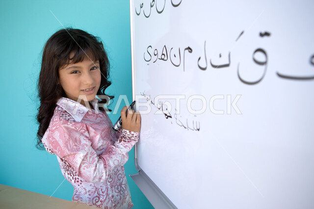 Enjoying learning the Arabic language, developing students' linguistic abilities, teaching students to form words in the Arabic language, a close-up portrait of a smiling Gulf Emirati Arab girl looking at the camera and holding a whiteboard pen in her han