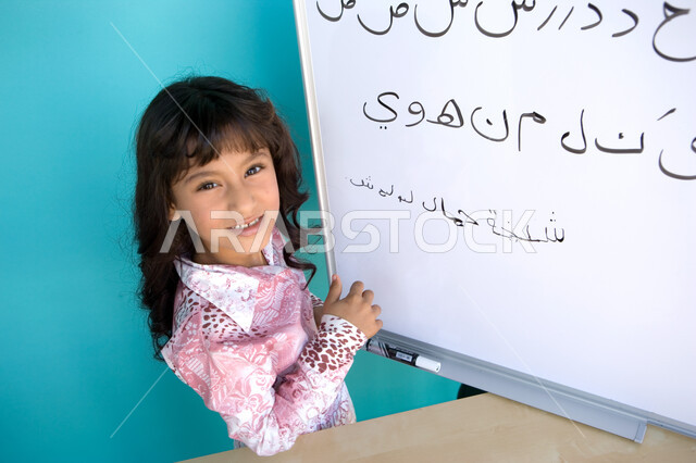الاستمتاع بتعلم اللغة العربية, تنمية القدرات اللغوية للطلاب, تعليم التلاميذ تكوين الكلمات باللغة العربية، بورتريه مقرب من الجانب لفتاة عربية خليجية إماراتية مبتسمة تنظر الى الكاميرا و تحمل بيدها قلم اللوح و تقوم بكتابة الكلمات على السبورة, خلفية زرقاء