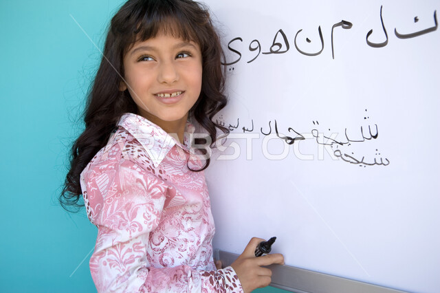 متعة حصة اللغة العربية, تنمية القدرات اللغوية والفكرية للطلاب, تعليم التلاميذ تكوين الكلمات باللغة العربية، بورتريه مقرب من الجانب لفتاة عربية خليجية إماراتية مبتسمة تنظر الى شيء ما و تحمل بيدها قلم اللوح و تقوم بكتابة الكلمات على السبورة, خلفية زرقاء