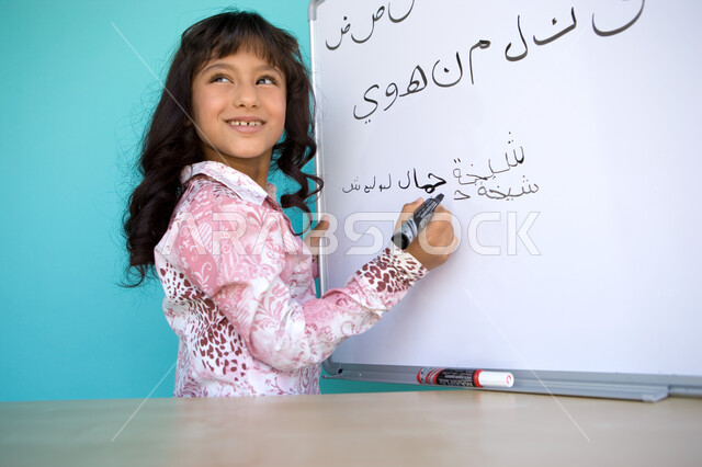 Fun Arabic language class, developing students’ linguistic and intellectual abilities, teaching students to form words in Arabic, close-up portrait from the side of a smiling Gulf Arab-Emirati girl looking at something, holding a blackboard pen in her han