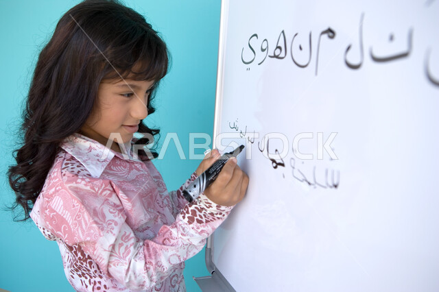 تنمية القدرات اللغوية والفكرية للطلاب, تعليم التلاميذ تكوين الكلمات باللغة العربية، جودة التعليم في الإمارات، بورتريه مقرب من الجانب لفتاة عربية خليجية إماراتية تحمل بيدها قلم اللوح و تقوم بكتابة الجمل على السبورة, متعة حصة اللغة العربية, خلفية زرقاء