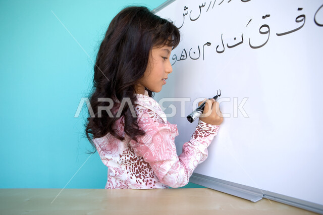 تنمية القدرات اللغوية والفكرية للطلاب, تعليم التلاميذ تكوين الكلمات باللغة العربية، جودة التعليم في الإمارات، بورتريه مقرب من الجانب لفتاة عربية خليجية إماراتية تحمل بيدها قلم اللوح و تقوم بكتابة الجمل على السبورة, متعة حصة اللغة العربية, خلفية زرقاء