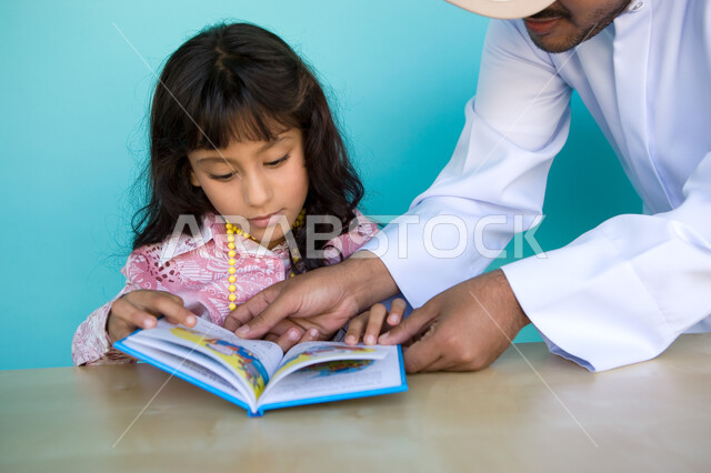 Encouraging children to learn and stop, gestures of attention and concentration while reading, close-up portrait of an Emirati Gulf Arab man wearing a ghutra and an Emirati kandura helping his daughter read, reading entertaining graphic stories, developin