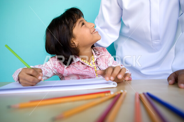 إيماءات السعادة والاستمتاع بمشاركة الأنشطة الفنية مع الأب، دور الأهل في تنمية المهارات, شغف وحب الرسم، بورتريه مقرب لفتاة صغيرة عربية خليجية إماراتية تقوم بالرسم والتلوين بالأقلام الخشبية رفقة والدها وتنظر اليه، رعاية المواهب والاهتمام بها , خلفية زرقاء